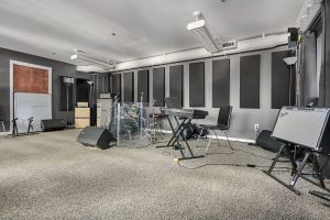 Studio room with drums, amps, keyboard, PA, and sound panels.