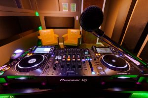 DJ Studio featuring Pioneer CDJs, color changing lights, automated recording, live-streaming capabilities, chairs, and microphones.